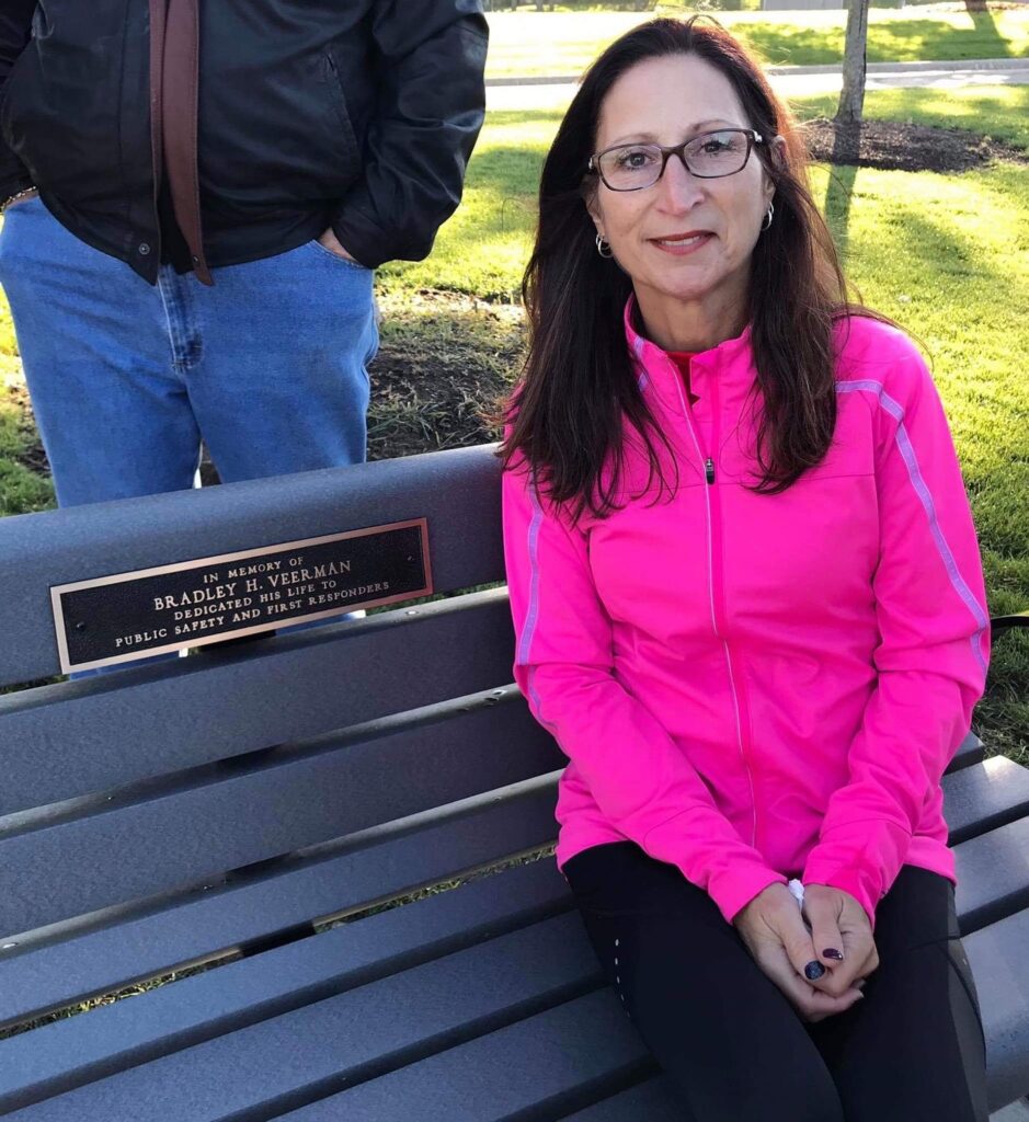 IMG_6636 April Veerman sits on a bench that honors her late husband, Brad, at Fireman's Park in New Lenox. She hopes people will "have a dollar" to support a 5k in his honor. (Photo by Stephanie Irvine)